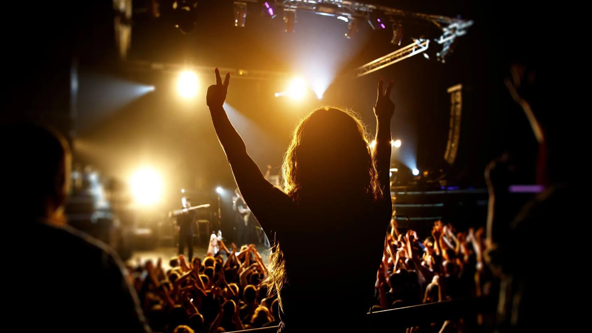 Pessoa com cabelos compridos e ondulados levanta os braços, formando um sinal de paz com cada mão, em meio a uma multidão num show, iluminada pelas luzes do palco. O público ao redor tem os braços levantados e é iluminado por holofotes laranjas.