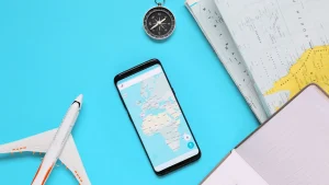 Objetos relacionados a viagens, como um pequeno avião de brinquedo, um smartphone mostrando um mapa, uma bússola e um caderno com uma caneta, dispostos sobre uma superfície azul clara. A imagem sugere planejamento ou preparação para uma viagem.