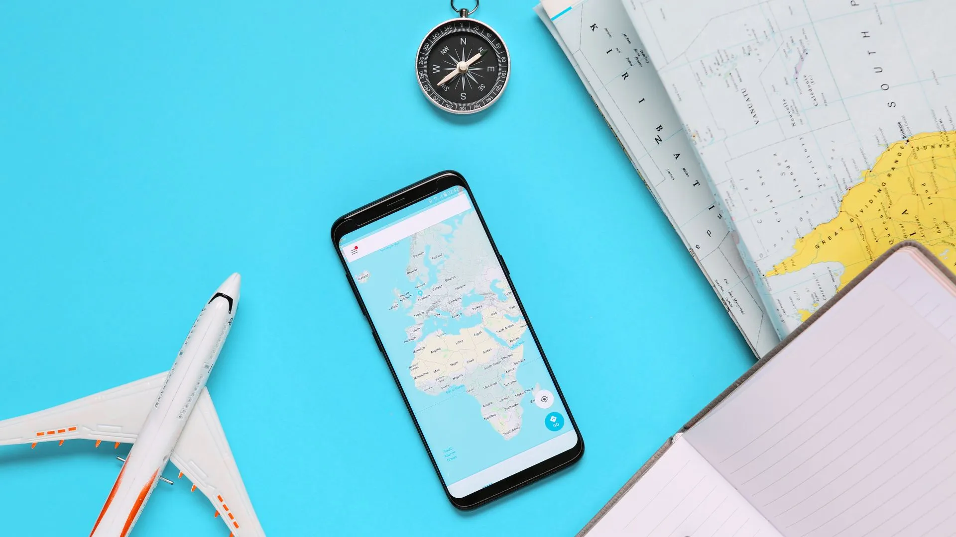 Objetos relacionados a viagens, como um pequeno avião de brinquedo, um smartphone mostrando um mapa, uma bússola e um caderno com uma caneta, dispostos sobre uma superfície azul clara. A imagem sugere planejamento ou preparação para uma viagem.