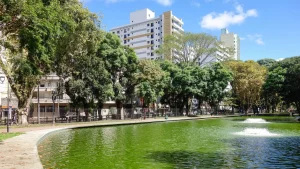 Vista do Passeio Público em Curitiba, Paraná, com um lago de água verde e uma fonte. A margem do lago é cercada por árvores frondosas, e prédios e edifícios altos podem ser vistos ao fundo, com um céu azul e poucas nuvens acima.