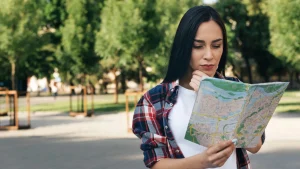 Uma foto externa mostra uma jovem mulher de cabelos escuros longos no centro, usando uma camiseta branca e uma camisa xadrez por cima, segurando e examinando um mapa dobrado. Ela tem uma expressão concentrada, com a mão direita no queixo. O fundo é um caminho pavimentado em um parque ou área verde, com árvores frondosas. A luz do sol está filtrada pelas árvores.