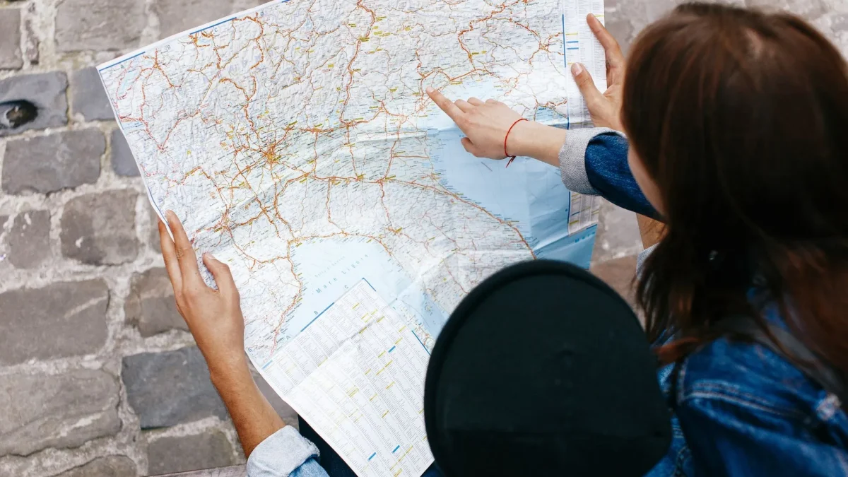 Duas pessoas estão examinando um mapa de papel detalhado. Uma delas, vestindo uma jaqueta jeans, está apontando para uma área específica no mapa, enquanto a outra pessoa segura o mapa. A cena é vista de cima, com o mapa ocupando a maior parte do quadro. As mãos e os braços das pessoas são visíveis, assim como o chão de paralelepípedos. A imagem sugere um momento de planejamento de viagem ou orientação.