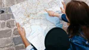 Duas pessoas estão examinando um mapa de papel detalhado. Uma delas, vestindo uma jaqueta jeans, está apontando para uma área específica no mapa, enquanto a outra pessoa segura o mapa. A cena é vista de cima, com o mapa ocupando a maior parte do quadro. As mãos e os braços das pessoas são visíveis, assim como o chão de paralelepípedos. A imagem sugere um momento de planejamento de viagem ou orientação.