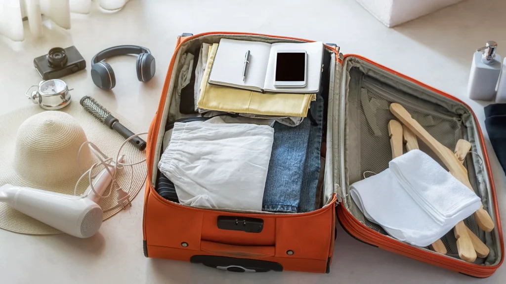 Mala de viagem de cor laranja aberta, recheada com roupas dobradas (shorts brancos, jeans) e itens pessoais, como um caderno ou bloco de anotações, uma caneta e um celular. Ao redor da mala, sobre um piso claro, estão dispostos outros acessórios essenciais para viagem: um chapéu de palha de aba larga, um secador de cabelo, uma escova de cabelo, fones de ouvido, uma câmera fotográfica e um pequeno recipiente de metal. No lado direito da mala, toalhas dobradas, cabides de madeira e frascos de amenities (sabonete líquido, shampoo, etc.) completam a cena.