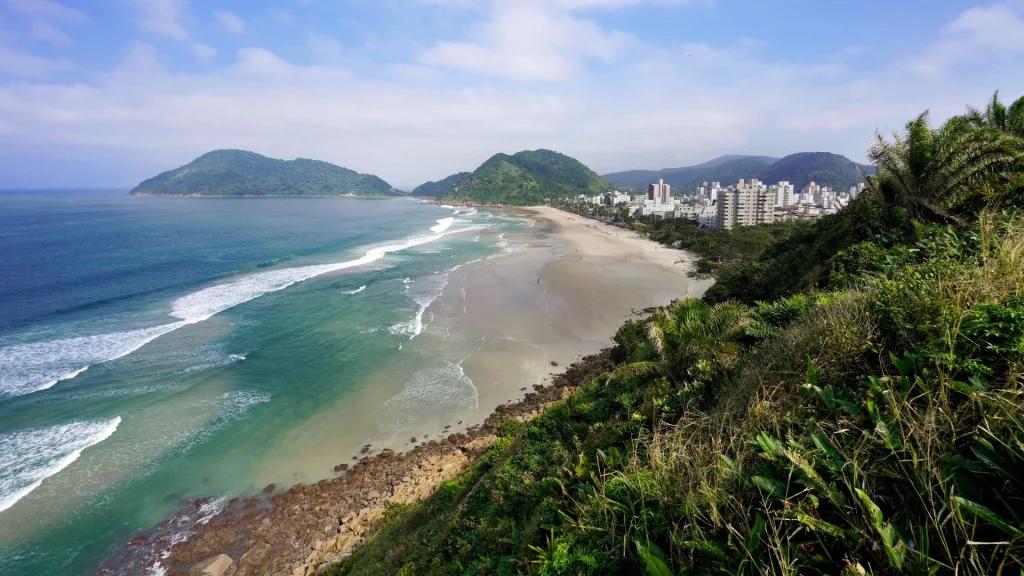 Vista panorâmica da praia, com vegetação nativa em primeiro plano, mar degradê entre azul e verde e montanhas ao fundo.