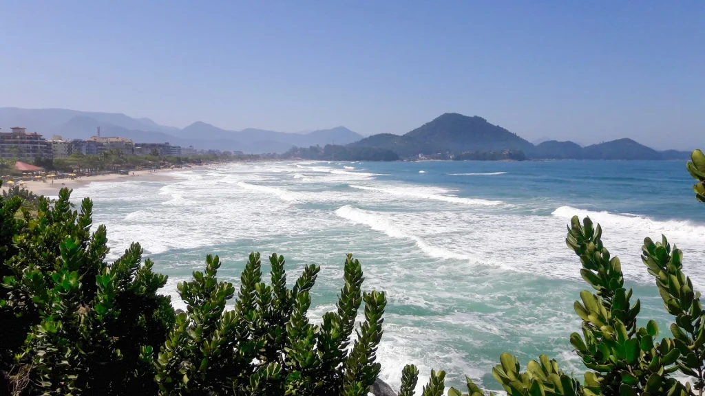 Vista panorâmica da Praia Grande em Ubatuba sob um céu azul. À esquerda, o mar verde-esmeralda com ondas brancas. No centro, uma longa faixa de areia clara com banhistas. À direita, um paredão de prédios residenciais modernos e vegetação tropical em primeiro plano.
