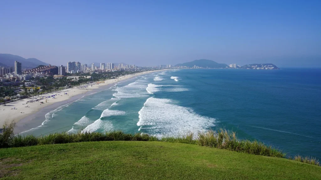 Vista panorâmica da Praia da Enseada a partir de um mirante gramado. O mar azul apresenta ondas longas e brancas que chegam a uma extensa faixa de areia clara. À esquerda, a orla é contornada por diversos prédios e montanhas ao fundo sob um céu limpo.