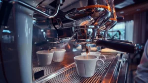 Um close-up em uma máquina de café expresso brilhante de cafeteria, mostrando a bandeja de metal e o cabeçote (grupo). Uma xícara de cerâmica branca está posicionada sob o bico de extração, com outras duas xícaras ao fundo, prontas para o preparo do café.