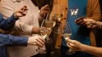 Quatro pessoas em um brinde, erguendo taças de vinho branco ou champanhe em primeiro plano. Eles seguram velas de faísca (sparklers) com luzes brilhantes, indicando uma celebração, provavelmente de ano novo ou aniversário.
