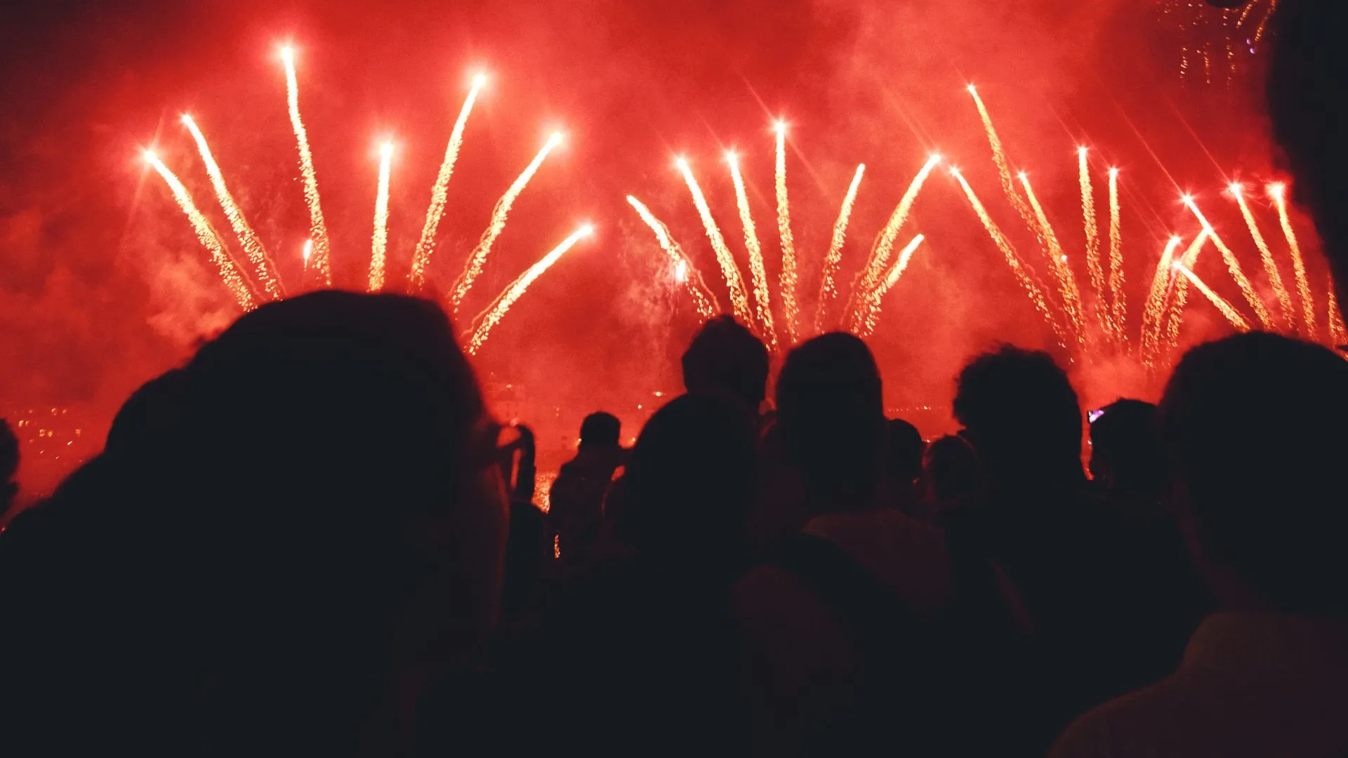 Exibição noturna intensa de fogos de artifício em tons de vermelho e dourado, com algumas explosões em formato de palmeira. A cena é vista por trás de uma multidão silhuetada, com balões de papel pendurados em um cordão sobre as cabeças das pessoas, criando um ambiente de celebração.