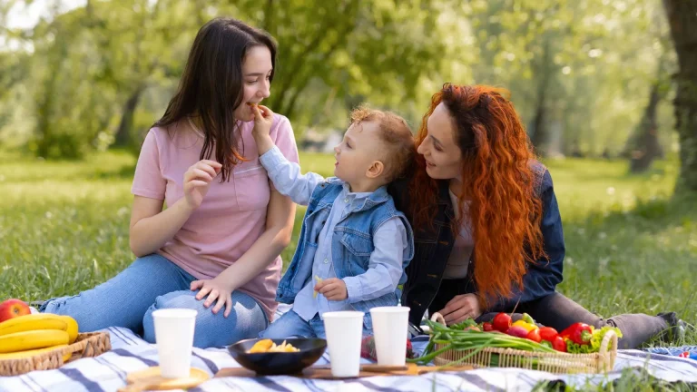Duas mulheres e uma criança pequena estão fazendo um piquenique em um parque em um dia ensolarado. A mulher da esquerda, de cabelo escuro e camiseta rosa, está sorrindo enquanto a criança, no centro, de colete jeans azul, estende a mão para lhe dar um pedaço de comida. A mulher da direita, de cabelo ruivo vibrante e jaqueta jeans, olha para a criança com um sorriso. Elas estão sentadas em um cobertor xadrez com frutas, vegetais e copos de papel na frente delas, cercadas por grama verde e árvores desfocadas.