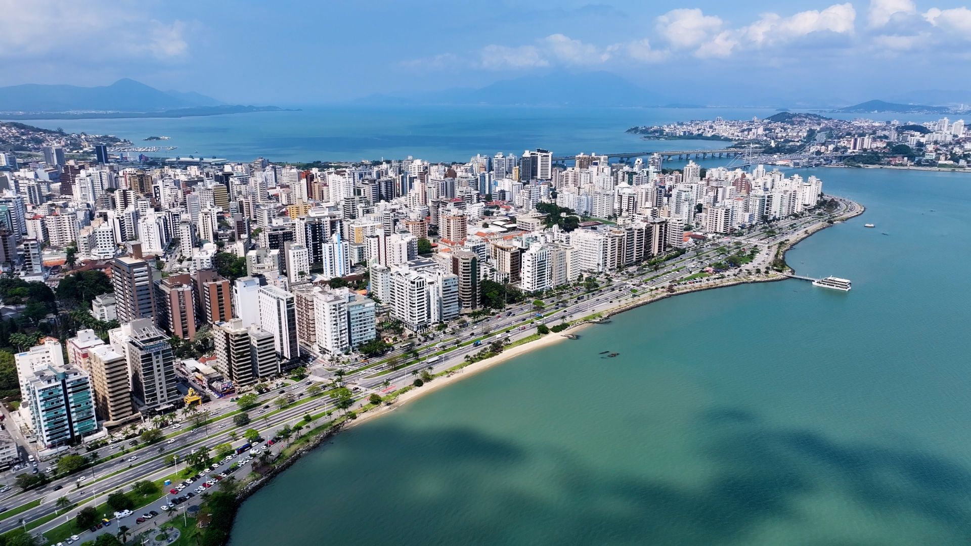 Vista aérea e panorâmica do horizonte de Florianópolis, Santa Catarina, Brasil. A imagem mostra uma densa área urbana de prédios de diversos tamanhos e cores claras, que se estende ao longo de uma ampla avenida costeira. A avenida é ladeada por uma faixa de areia estreita e pelo mar, com águas em tons de azul-turquesa e verde-azulado. Ao fundo, pode-se ver o restante da cidade, colinas cobertas de vegetação, e uma ponte que liga a ilha ao continente. O céu é azul com algumas nuvens brancas.