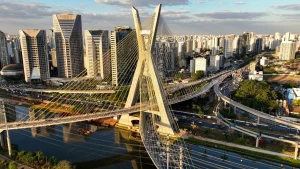 Vista aérea da Ponte Octávio Frias de Oliveira (Ponte Estaiada) em São Paulo, Brasil, destacando sua estrutura de cabos em 'X' e o mastro central, em contraste com a paisagem urbana circundante e os arranha-céus.
