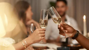 Duas pessoas fazem um brinde com taças de champanhe durante uma celebração. O foco está nas taças e nas mãos, com luzes e velas no fundo.