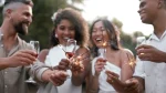 Um grupo de quatro jovens adultos, dois homens e duas mulheres, sorrindo e celebrando ao ar livre. Eles seguram taças com uma bebida avermelhada (possivelmente vinho ou espumante) e pequenos fogos de artifício (sparklers) acesos, que estão soltando faíscas. As mulheres estão vestindo roupas brancas, e os homens usam camisas claras. A cena transmite alegria e celebração, sugerindo uma festa ou um evento especial, como o Ano Novo.
