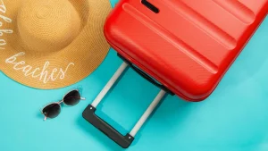Mala de viagem rígida vermelha com a alça estendida, óculos de sol de armação rosa e dourada e um chapéu de palha com a palavra 'beaches' (praias) bordada em branco, tudo disposto sobre um fundo azul-turquesa. O cenário sugere férias de verão e viagem.