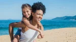 Mãe e filha sorrindo e se divertindo na praia. A mãe, de cabelo cacheado, está carregando a filha nas costas. A filha, que sorri e olha para frente, está com os braços ao redor do pescoço da mãe. O fundo é uma paisagem de praia com areia dourada, mar azul e morros ao longe.