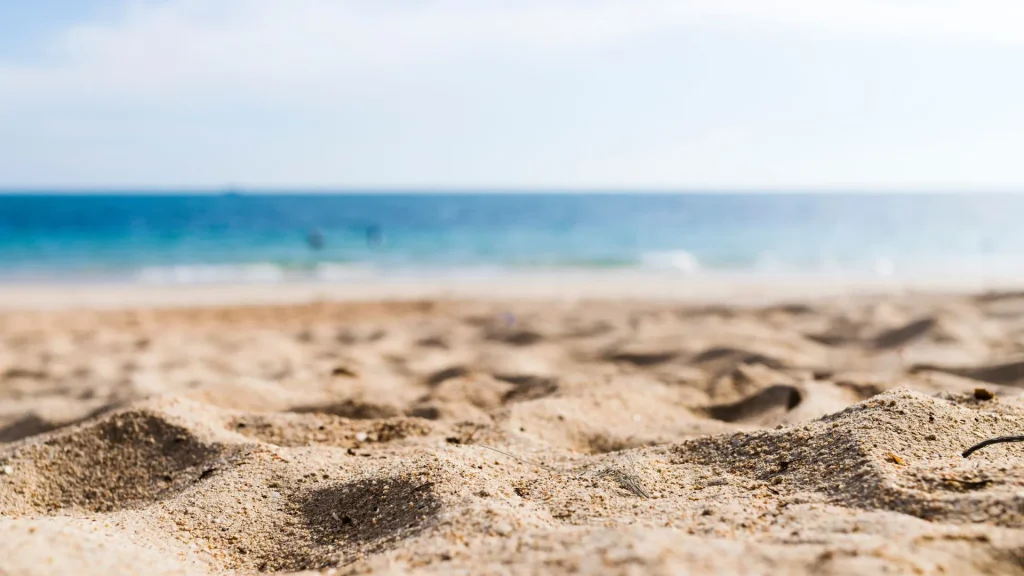 Vista em close-up de uma praia de areia clara, com o foco na textura granulada da areia em primeiro plano. O horizonte do oceano, com águas azuis e claras sob um céu claro, está desfocado no fundo.