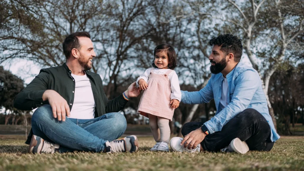 Uma família composta por dois pais e uma filha pequena sentada em um gramado de um parque. Os dois homens estão sentados de pernas cruzadas no chão, sorrindo e olhando para a criança que está no centro, de pé, usando um vestido rosa. O homem à esquerda veste uma camisa branca sob uma jaqueta verde escura. O homem à direita veste uma camisa social azul clara e tem barba. O fundo é de árvores desfocadas.