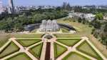Vista aérea do Jardim Botânico de Curitiba, Paraná. No centro, destaca-se a icônica estufa de metal e vidro em estilo art nouveau com três cúpulas. À frente da estufa, estende-se um jardim em estilo francês com gramados simétricos em formatos geométricos, cercados por sebes verdes e caminhos pavimentados. Ao fundo, uma densa área de mata nativa e a silhueta urbana da cidade sob um céu azul claro.