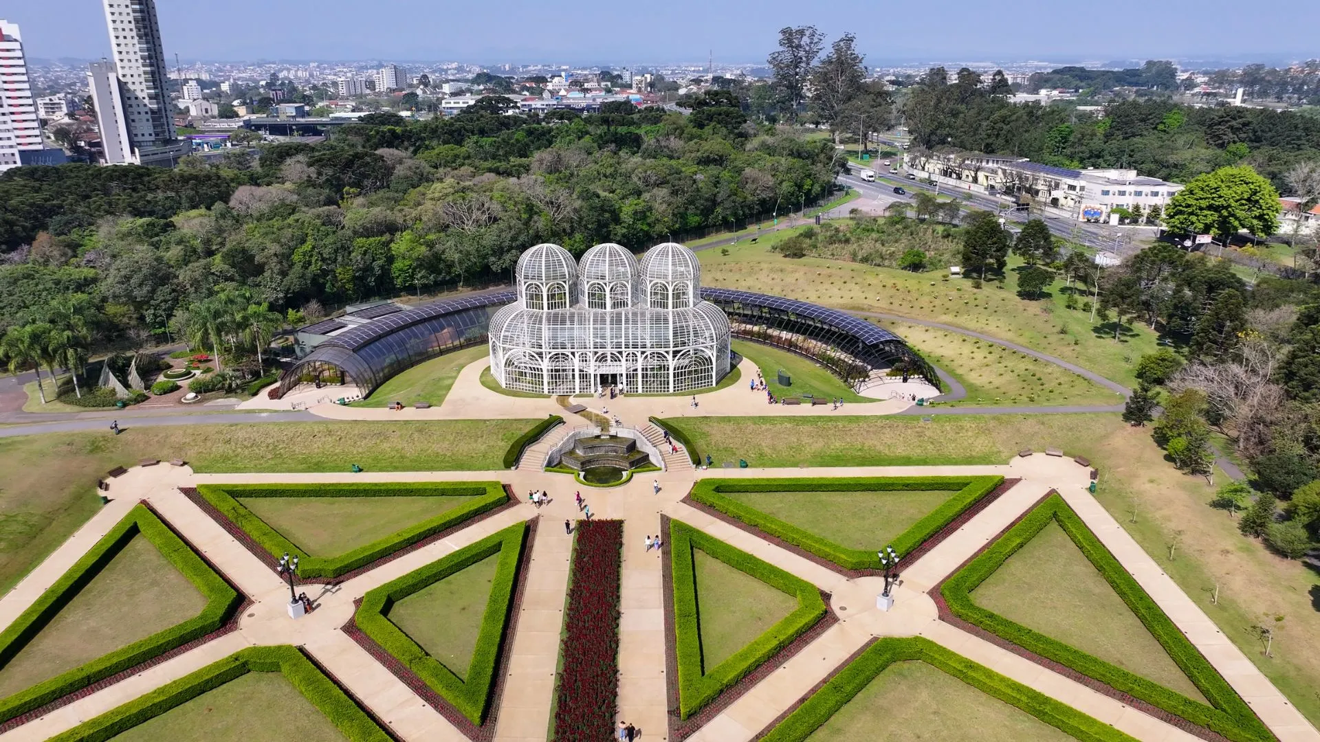 Vista aérea do Jardim Botânico de Curitiba, Paraná. No centro, destaca-se a icônica estufa de metal e vidro em estilo art nouveau com três cúpulas. À frente da estufa, estende-se um jardim em estilo francês com gramados simétricos em formatos geométricos, cercados por sebes verdes e caminhos pavimentados. Ao fundo, uma densa área de mata nativa e a silhueta urbana da cidade sob um céu azul claro.