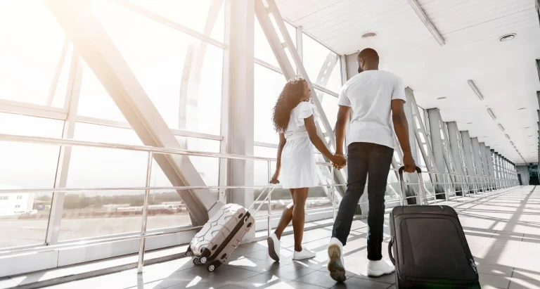 Um casal negro de costas, de mãos dadas, caminha por um corredor amplo e iluminado de um aeroporto. A mulher, à esquerda, usa um vestido branco curto e puxa uma mala de rodinhas bege. O homem, à direita, veste camiseta branca e calça jeans escura, carregando uma mala preta. Eles olham um para o outro enquanto caminham em direção a uma grande janela de vidro que revela uma estrutura metálica externa sob a luz do sol.