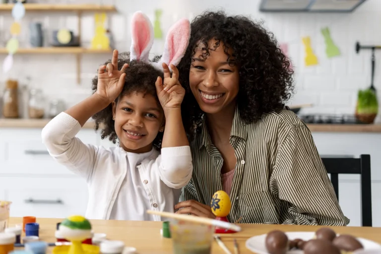 Uma mulher e uma criança negra sorriem alegremente em uma cozinha decorada para a Páscoa. A criança, à esquerda, usa uma tiara de orelhas de coelho rosa e brancas, enquanto faz um gesto de "orelhas" com as mãos sobre a cabeça. A mulher, à direita, está abraçada à criança. À frente delas, sobre uma mesa, há um ovo de Páscoa amarelo decorado. Ao fundo, a cozinha branca exibe enfeites coloridos de coelhinhos pendurados.