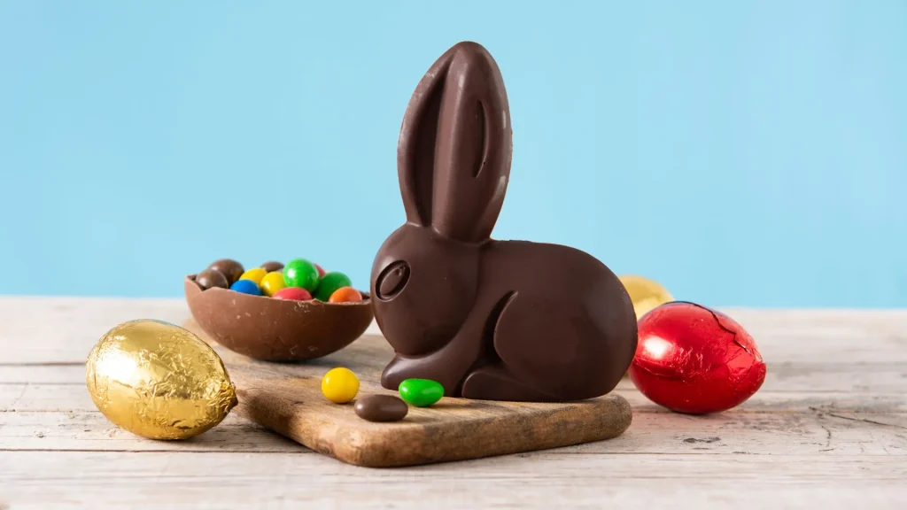 Um coelho de chocolate amargo posicionado no centro de uma tábua de madeira clara. Ao lado dele, há uma metade de ovo de chocolate recheada com confeitos coloridos, além de ovos de Páscoa menores embrulhados em papel alumínio dourado e vermelho. Alguns confeitos estão espalhados pela superfície. O fundo é de um azul sólido e suave, criando um contraste festivo e clean.