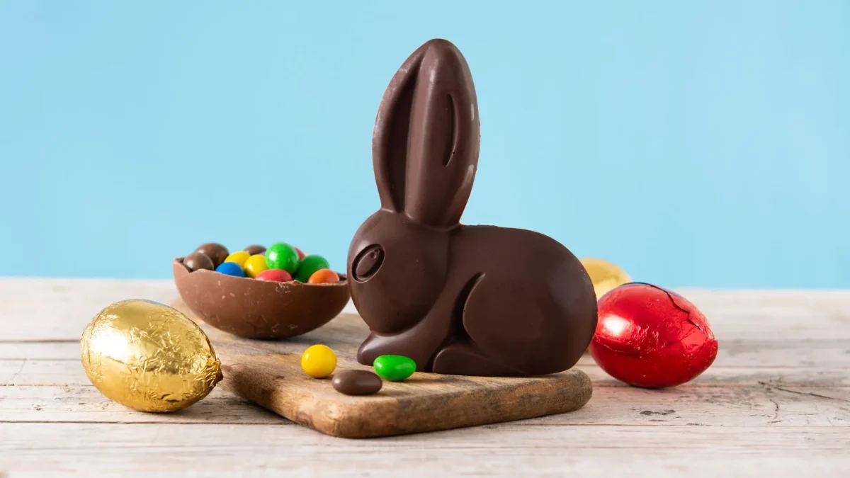 Um coelho de chocolate amargo posicionado no centro de uma tábua de madeira clara. Ao lado dele, há uma metade de ovo de chocolate recheada com confeitos coloridos, além de ovos de Páscoa menores embrulhados em papel alumínio dourado e vermelho. Alguns confeitos estão espalhados pela superfície. O fundo é de um azul sólido e suave, criando um contraste festivo e clean.