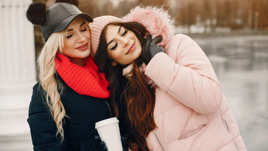 Duas mulheres jovens em um clima carinho em um ambiente externo de inverno. A mulher à esquerda tem cabelos loiros longos e ondulados, usa um boné de couro preto com pompons, um cachecol vermelho vibrante e uma jaqueta azul-escura. Ela está com os olhos fechados e encosta o rosto suavemente na cabeça da amiga. A mulher à direita tem cabelos castanhos longos, usa uma touca de lã rosa claro e uma jaqueta puffer rosa com capuz de pele sintética. Ela também está com os olhos fechados, sorrindo levemente e segurando um copo térmico branco. O fundo está desfocado, sugerindo um parque ou área aberta durante o dia.