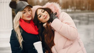 Duas mulheres jovens em um clima carinho em um ambiente externo de inverno. A mulher à esquerda tem cabelos loiros longos e ondulados, usa um boné de couro preto com pompons, um cachecol vermelho vibrante e uma jaqueta azul-escura. Ela está com os olhos fechados e encosta o rosto suavemente na cabeça da amiga. A mulher à direita tem cabelos castanhos longos, usa uma touca de lã rosa claro e uma jaqueta puffer rosa com capuz de pele sintética. Ela também está com os olhos fechados, sorrindo levemente e segurando um copo térmico branco. O fundo está desfocado, sugerindo um parque ou área aberta durante o dia.