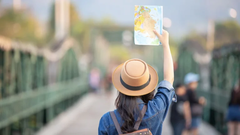 Vista de costas de uma mulher com chapéu e jaqueta jeans, segurando um mapa aberto acima da cabeça enquanto caminha sobre uma ponte de estrutura metálica verde. Ao fundo, o cenário está desfocado com outras pessoas e montanhas sob a luz do dia.