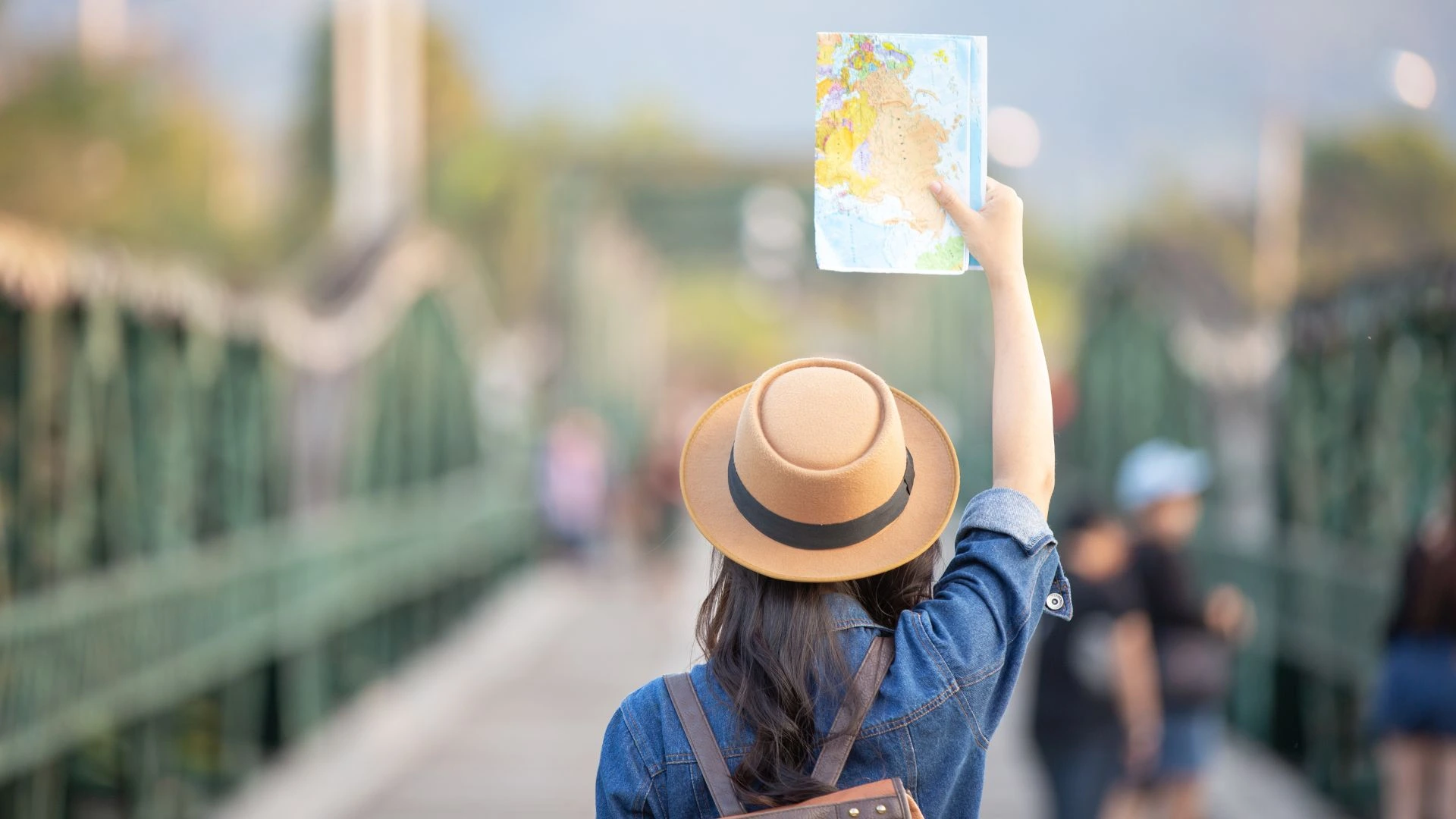 Vista de costas de uma mulher com chapéu e jaqueta jeans, segurando um mapa aberto acima da cabeça enquanto caminha sobre uma ponte de estrutura metálica verde. Ao fundo, o cenário está desfocado com outras pessoas e montanhas sob a luz do dia.