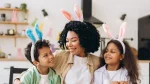 Uma mulher negra sorridente abraça duas crianças (um menino e uma menina) em uma cozinha decorada para a Páscoa. Todos os três usam tiaras com orelhas de coelho: as da mulher e da menina são cor-de-rosa, enquanto as do menino são azuis. As crianças olham para a mulher com carinho, e o ambiente ao fundo está iluminado, com coelhos de papel coloridos enfeitando as prateleiras.