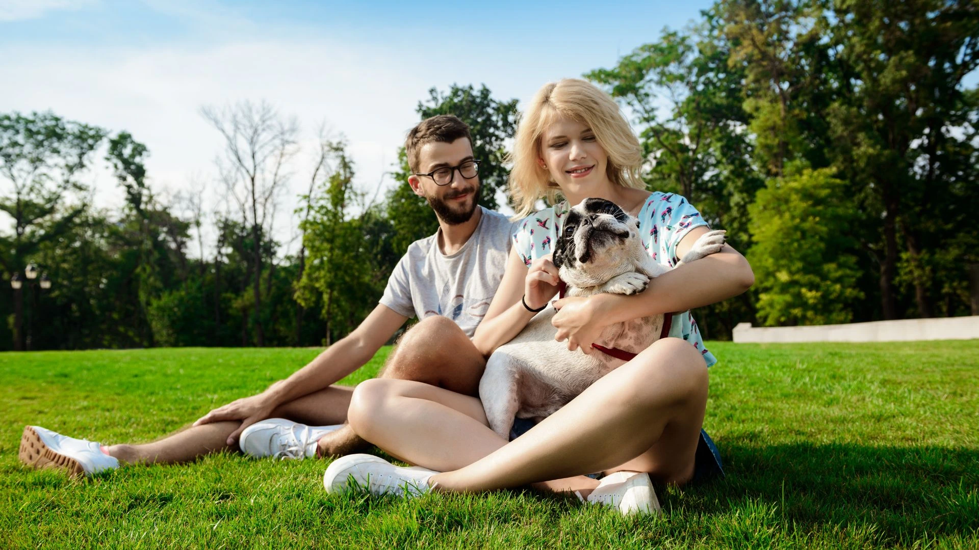 Um casal jovem — um homem de óculos e uma mulher loira — sorri enquanto descansa em um gramado ensolarado de um parque, segurando carinhosamente um cachorro da raça buldog francês no colo. O ambiente é cercado por árvores altas e folhagens verdes sob um céu claro.