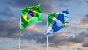 Bandeiras do Brasil e de Mato Grosso do Sul hasteadas em mastros metálicos contra um céu com nuvens em tons de azul e laranja.