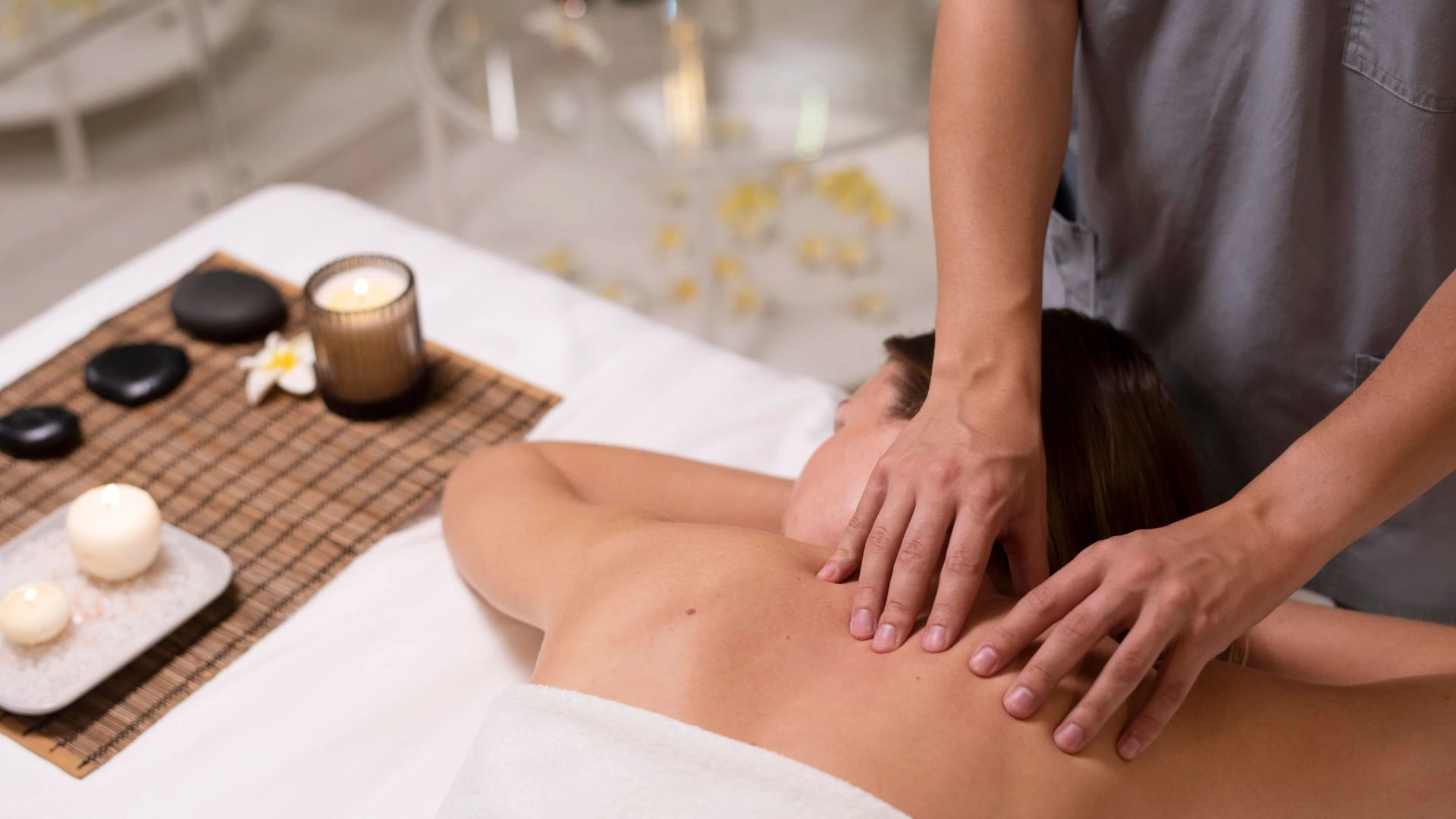 A imagem mostra uma pessoa recebendo uma massagem relaxante nas costas em um ambiente de spa. Ao lado da maca, há uma composição zen com pedras vulcânicas (hot stones), velas acesas que proporcionam uma iluminação suave e acolhedora, e uma flor de jasmim, reforçando a atmosfera de tranquilidade e autocuidado. As mãos do terapeuta aplicam pressão suave, simbolizando o alívio de tensões e o foco no bem-estar físico e mental.