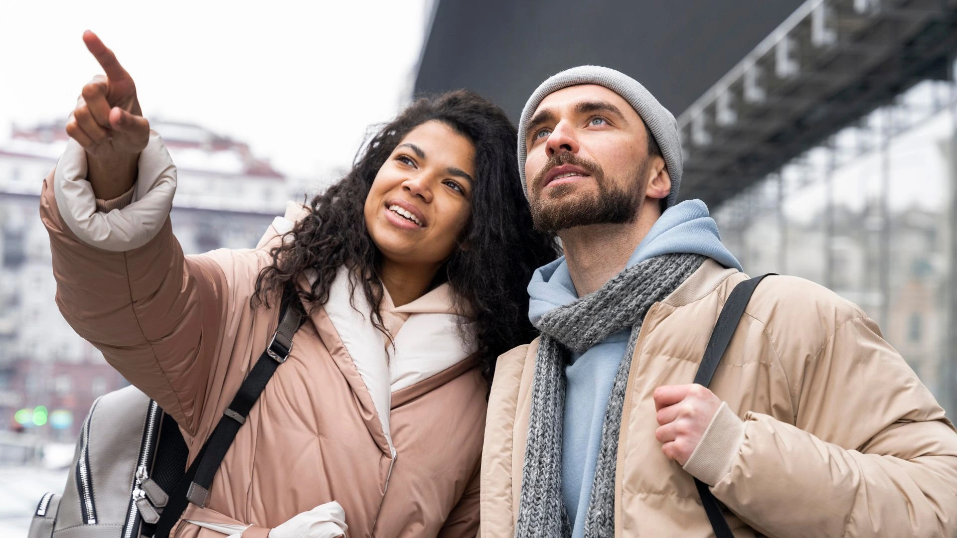 Uma fotografia de plano médio de um casal jovem em um ambiente urbano durante o inverno. À esquerda, uma mulher negra com cabelos cacheados e volumosos sorri enquanto aponta para algo fora do quadro. À direita, um homem branco com barba, vestindo uma touca cinza e cachecol, olha atentamente na mesma direção. Ambos usam casacos acolchoados em tons de bege e carregam mochilas, sugerindo que são turistas explorando a cidade. O fundo está levemente desfocado, mostrando estruturas arquitetônicas e um céu nublado.