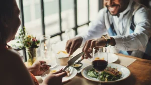 Casal negro sorridente desfruta de um jantar romântico em um restaurante elegante. O homem, em foco no lado direito, usa um relógio dourado e corta o alimento em seu prato, enquanto a mulher, em primeiro plano à esquerda (de costas), segura um garfo. Sobre a mesa de madeira, há taças de vinho tinto, pratos com peixe grelhado e salada, além de um pequeno arranjo de flores. A iluminação é suave, vindo de grandes janelas ao fundo.