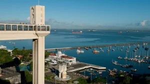 Vista panorâmica do Elevador Lacerda em Salvador, Bahia, com destaque para a Baía de Todos-os-Santos ao fundo, barcos ancorados no porto e o Mercado Modelo à beira-mar sob um céu azul.