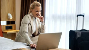 Mulher loira com traje executivo bege sorri enquanto utiliza um laptop sentada em uma cama de hotel. Ao fundo, uma janela com cortinas claras e uma mala de viagem preta, ilustrando o conceito de trabalho remoto ou viagem corporativa.