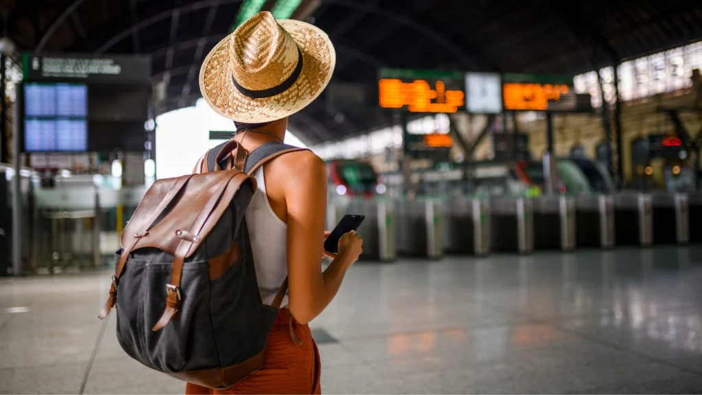 Vista de costas de uma viajante em uma estação de trem. Ela usa um chapéu de palha, regata branca, calça laranja e uma mochila de lona escura com detalhes em couro marrom. A mulher segura um celular enquanto observa o painel eletrônico de horários ao fundo, que está levemente desfocado. O ambiente é amplo, com arquitetura clássica de terminal ferroviário e catracas de acesso visíveis.
