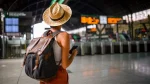 Vista de costas de uma viajante em uma estação de trem. Ela usa um chapéu de palha, regata branca, calça laranja e uma mochila de lona escura com detalhes em couro marrom. A mulher segura um celular enquanto observa o painel eletrônico de horários ao fundo, que está levemente desfocado. O ambiente é amplo, com arquitetura clássica de terminal ferroviário e catracas de acesso visíveis.