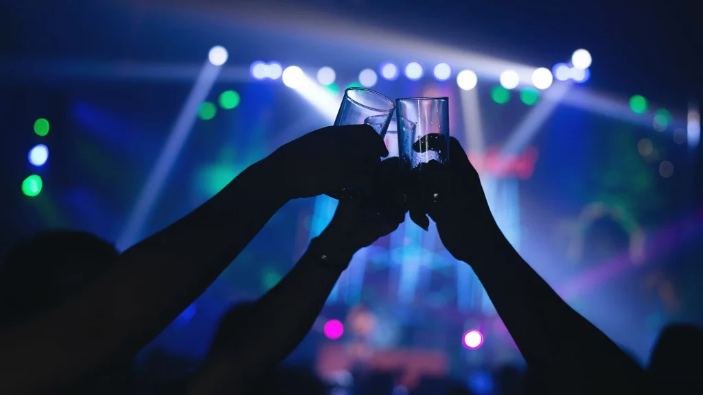 Uma silhueta em primeiro plano mostra três mãos erguendo copos de vidro para um brinde em um ambiente de festa ou show. O fundo está desfocado, iluminado por luzes de palco vibrantes em tons de azul, roxo e verde, criando uma atmosfera de vida noturna e celebração.