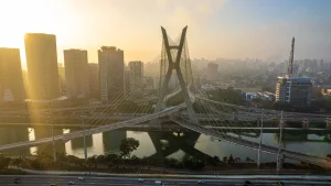 Vista aérea da Ponte Estaiada Octávio Frias de Oliveira em São Paulo, sob a luz dourada do nascer ou pôr do sol. A ponte, com seu característico mastro em formato de "X", cruza o Rio Pinheiros, conectando vias expressas movimentadas. Ao fundo, arranha-céus modernos e a silhueta da cidade estão envoltos em uma leve névoa matinal, enquanto o sol brilha intensamente no canto superior esquerdo, refletindo na água e nas fachadas de vidro dos prédios.
