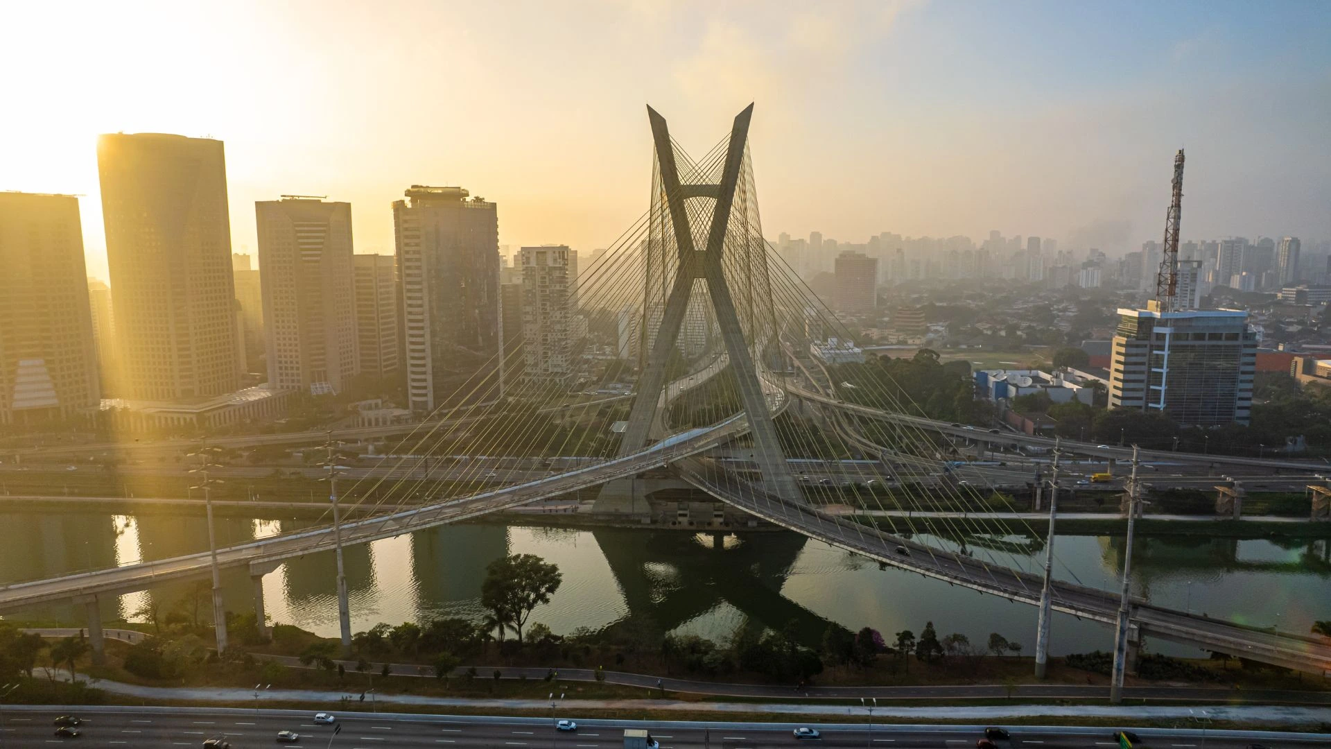 Vista aérea da Ponte Estaiada Octávio Frias de Oliveira em São Paulo, sob a luz dourada do nascer ou pôr do sol. A ponte, com seu característico mastro em formato de "X", cruza o Rio Pinheiros, conectando vias expressas movimentadas. Ao fundo, arranha-céus modernos e a silhueta da cidade estão envoltos em uma leve névoa matinal, enquanto o sol brilha intensamente no canto superior esquerdo, refletindo na água e nas fachadas de vidro dos prédios.