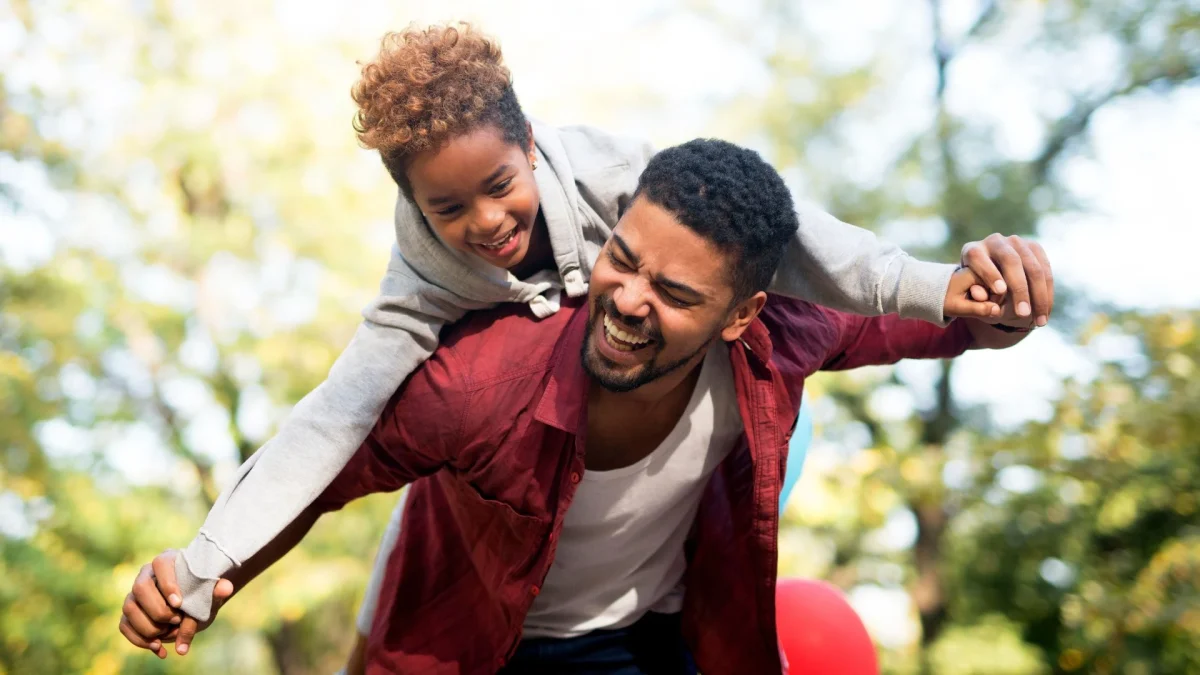 Um homem adulto negro, com barba curta e vestindo uma camisa de botão vinho sobre uma camiseta branca, carrega uma menina negra de cerca de 8 anos nas costas. Ambos estão rindo com expressão de grande alegria. A menina está deitada sobre as costas dele, com os braços abertos para os lados, enquanto o homem segura suas mãos, simulando as asas de um avião. Ela tem cabelos cacheados curtos e usa um moletom cinza. O fundo é um parque com árvores verdes levemente desfocadas pela luz do sol, criando uma atmosfera calorosa e descontraída. É possível ver o detalhe de balões coloridos na parte inferior da imagem.