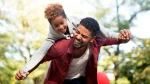 Um homem adulto negro, com barba curta e vestindo uma camisa de botão vinho sobre uma camiseta branca, carrega uma menina negra de cerca de 8 anos nas costas. Ambos estão rindo com expressão de grande alegria. A menina está deitada sobre as costas dele, com os braços abertos para os lados, enquanto o homem segura suas mãos, simulando as asas de um avião. Ela tem cabelos cacheados curtos e usa um moletom cinza. O fundo é um parque com árvores verdes levemente desfocadas pela luz do sol, criando uma atmosfera calorosa e descontraída. É possível ver o detalhe de balões coloridos na parte inferior da imagem.