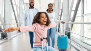 Uma menina negra de cabelos cacheados, vestindo uma blusa rosa, está sentada em sua mala e deslizando por um corredor de aeroporto com os braços abertos e uma expressão de pura alegria. Ao fundo, seus pais — um homem e uma mulher negros — caminham sorridentes, acompanhando a diversão da filha. O ambiente é moderno, com grandes janelas de vidro e estruturas metálicas, banhado por uma luz natural brilhante.