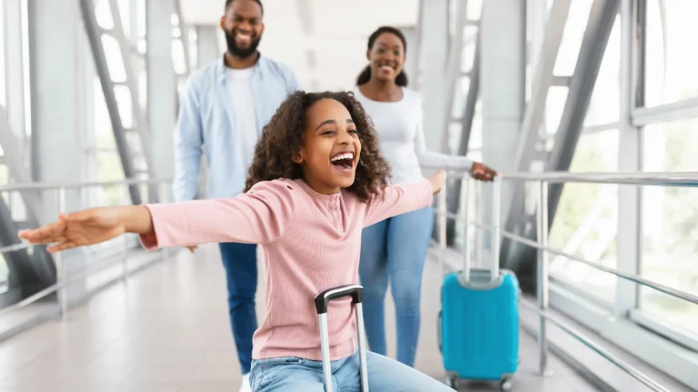 Uma menina negra de cabelos cacheados, vestindo uma blusa rosa, está sentada em sua mala e deslizando por um corredor de aeroporto com os braços abertos e uma expressão de pura alegria. Ao fundo, seus pais — um homem e uma mulher negros — caminham sorridentes, acompanhando a diversão da filha. O ambiente é moderno, com grandes janelas de vidro e estruturas metálicas, banhado por uma luz natural brilhante.